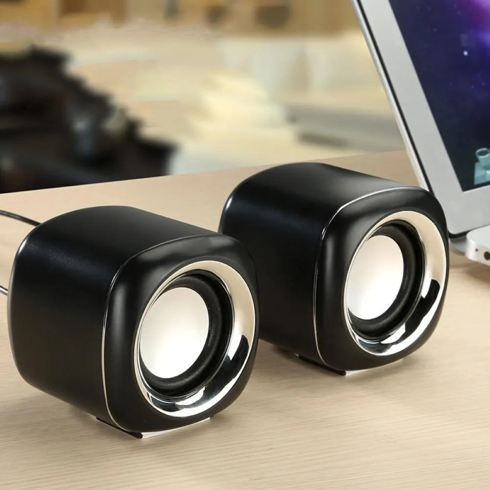 A pair of compact black wired desktop computer speakers with a volume scroll wheel, shown next to a 3.5mm audio plug.