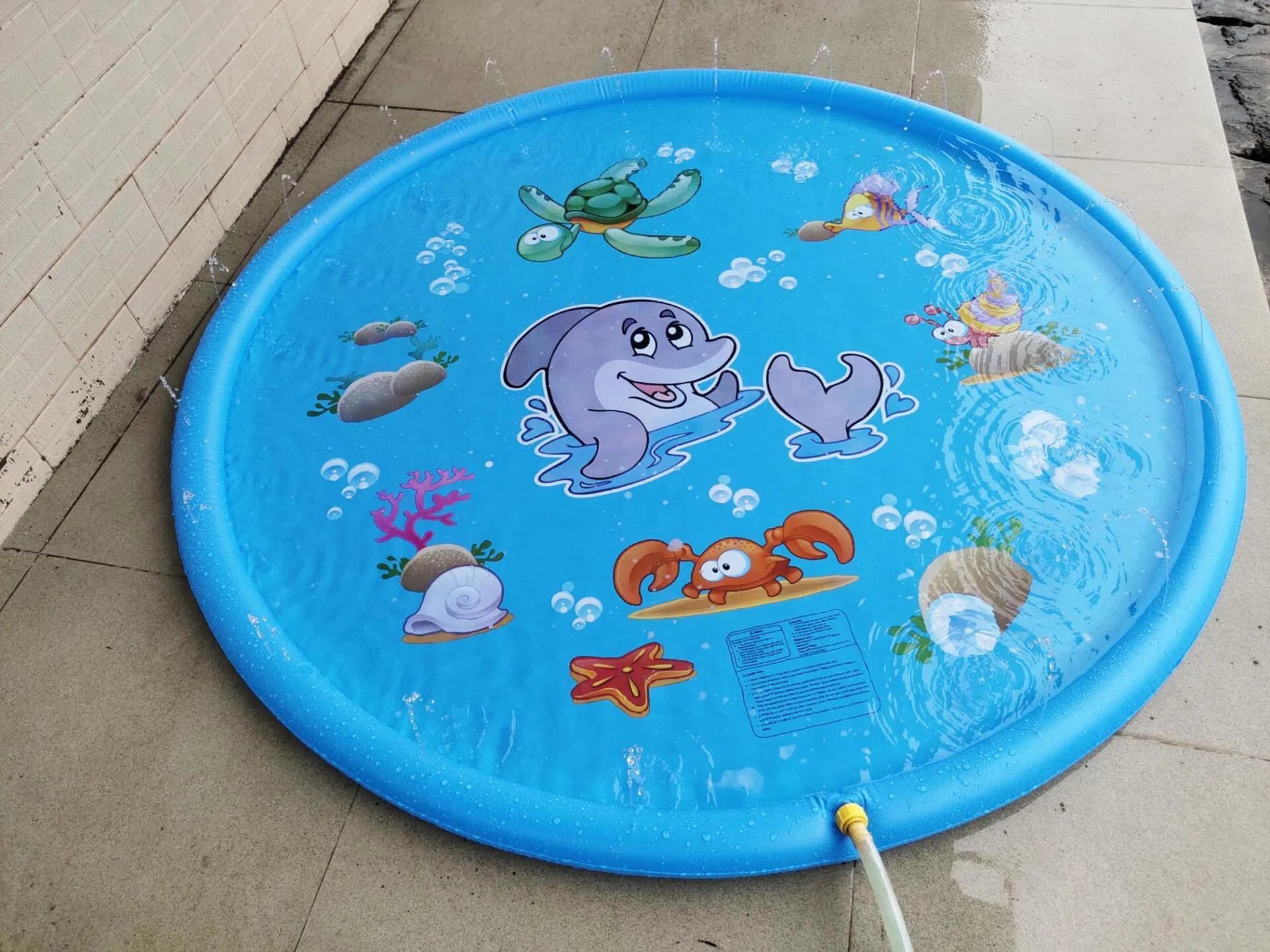 Blue PVC children's water spray play mat with marine animal designs, shown connected to a garden hose