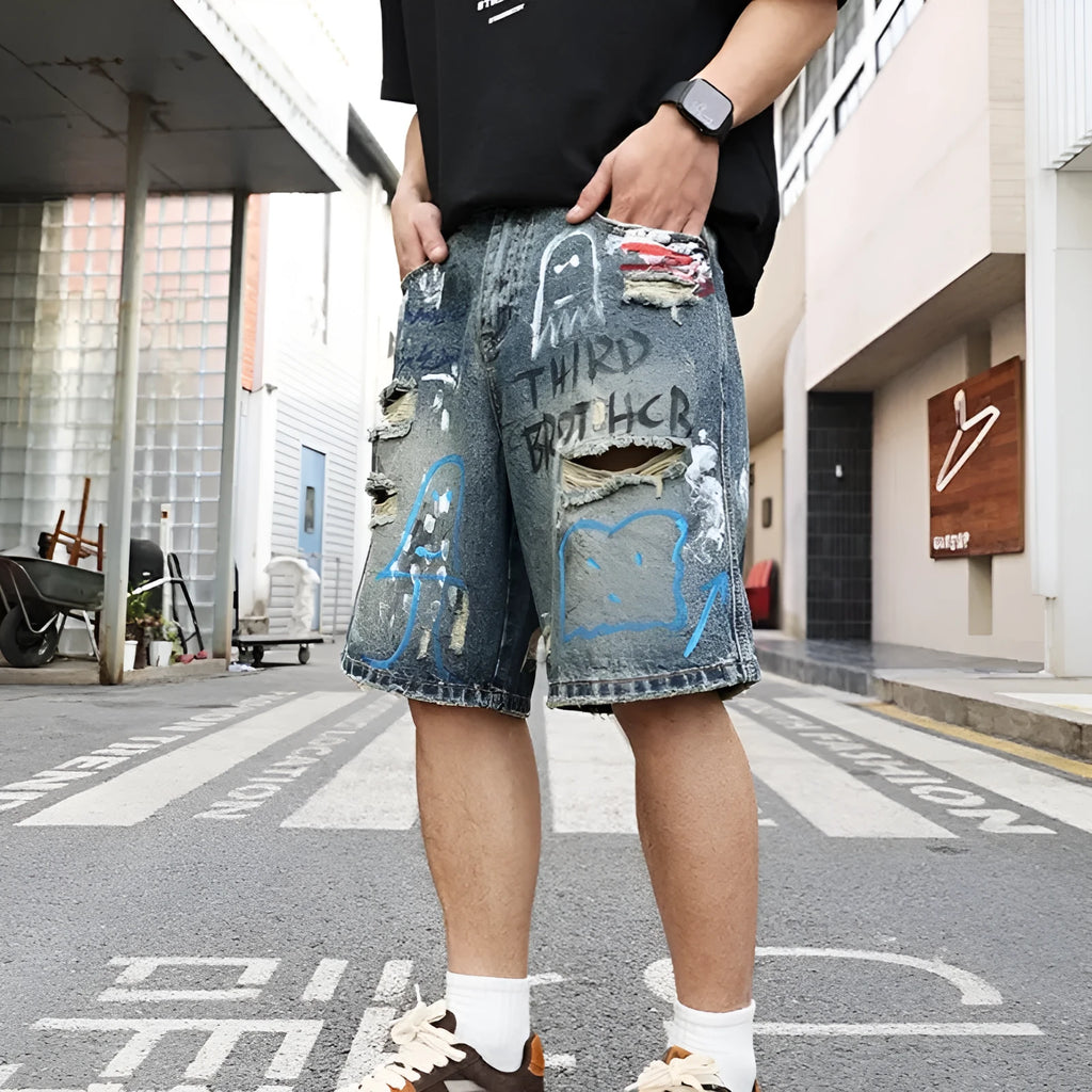 Blue men's denim shorts with ripped knees and graffiti detailing, showing straight leg cut and zipper fly.