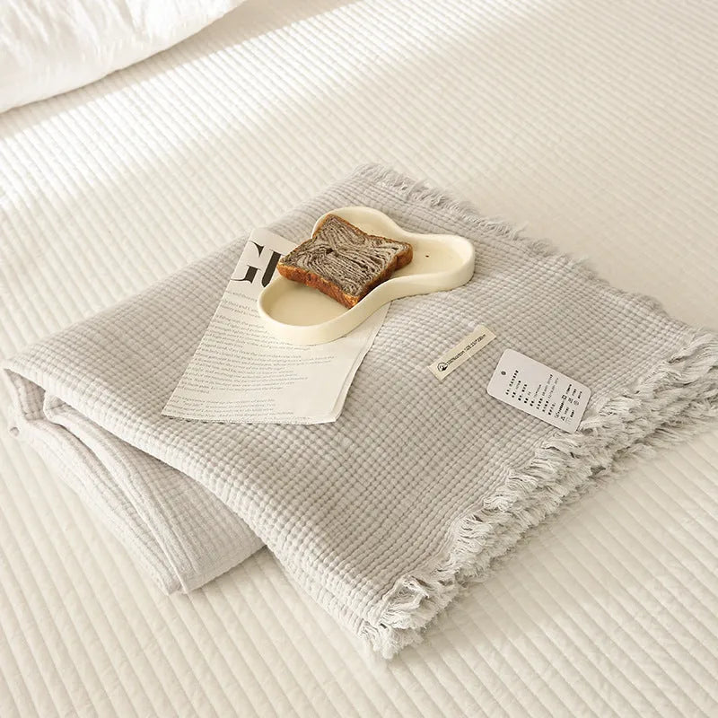 Cotton four-layer gauze blanket with tassel trim in various colours draped over a sofa.