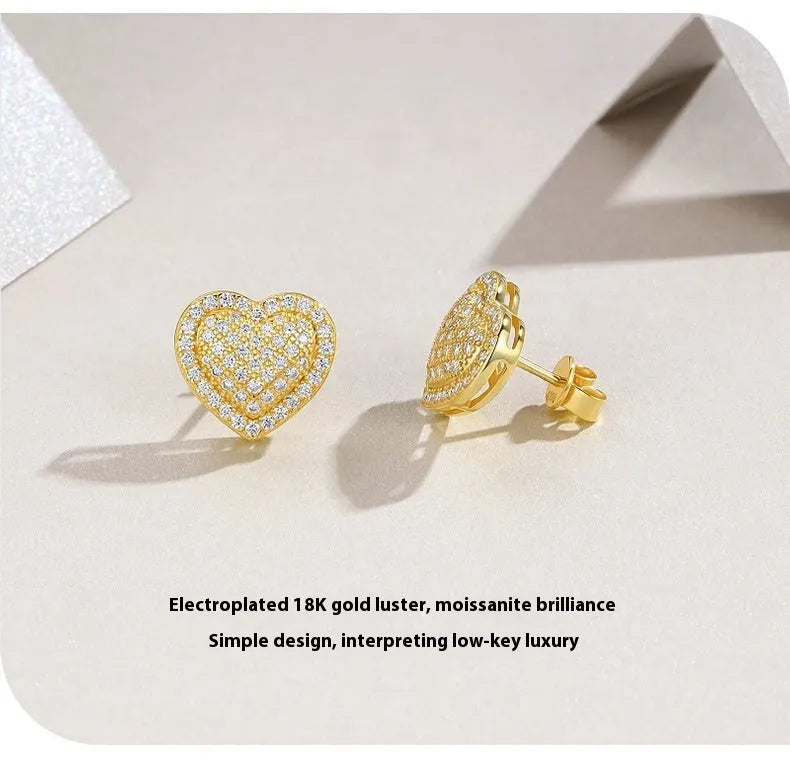 A pair of gold-colour heart-shaped stud earrings with a moissanite stone, on a plain background.