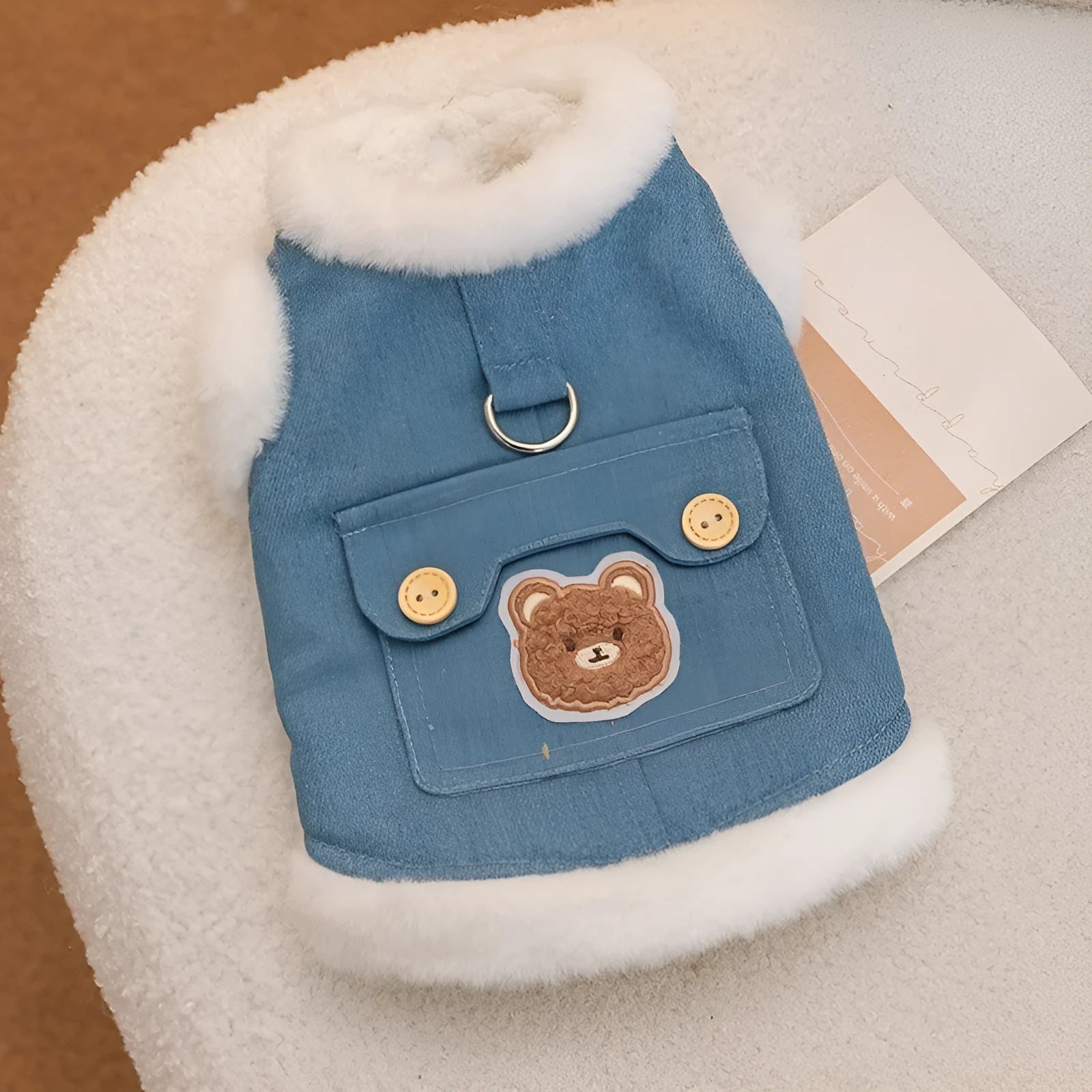 Denim dog vest with white wool lining, embroidered teddy bear patches, wooden buttons, and metal D-ring on blue background